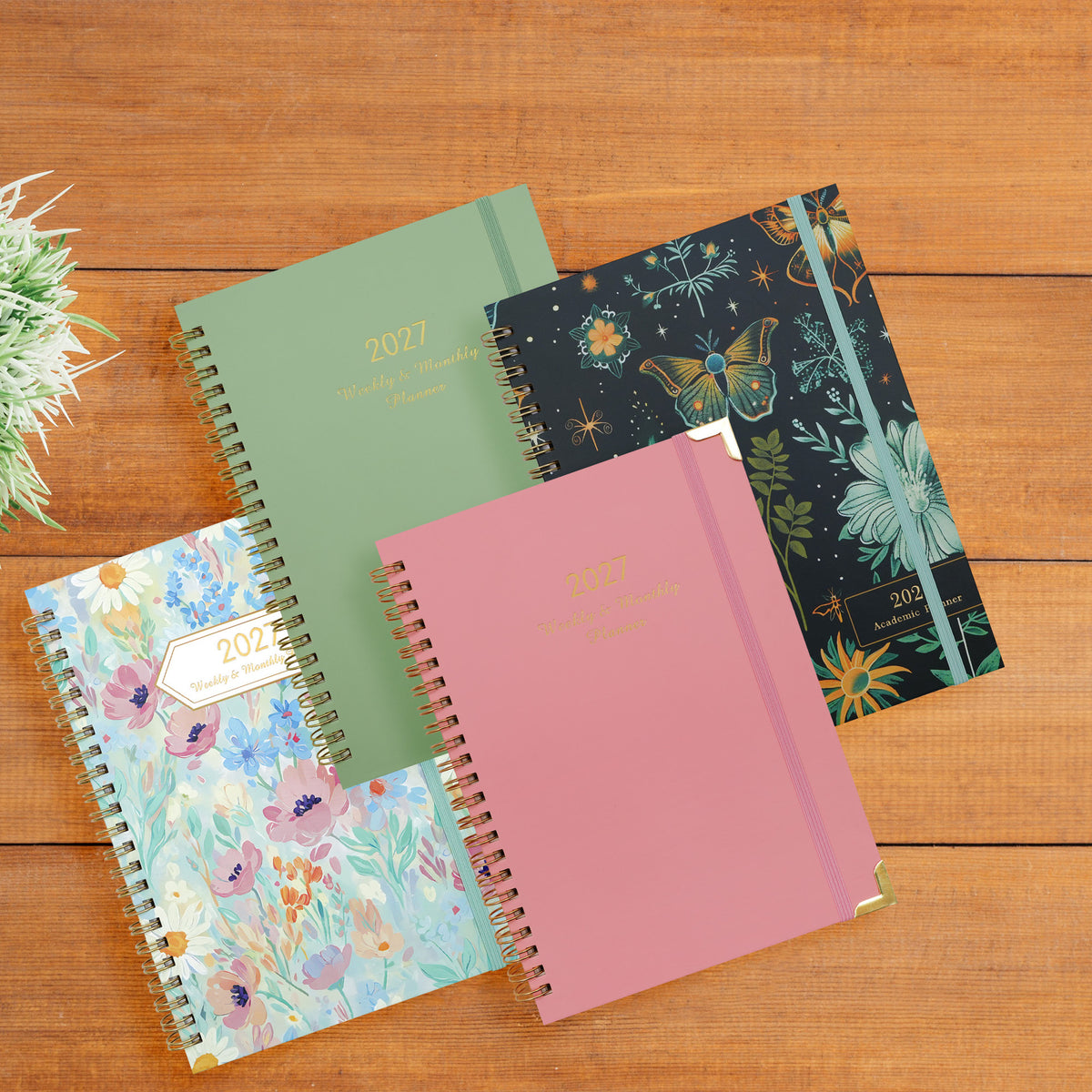 Four 2027 A5 spiral planners with pink, green, floral and dark botanical covers arranged on a wooden table